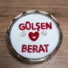 Kişiye özel isimli mumun üstten yakın çekimi: beyaz mum yüzeyinde kırmızı harflerle yazılmış isimler, kalp figürü ve iki fitil