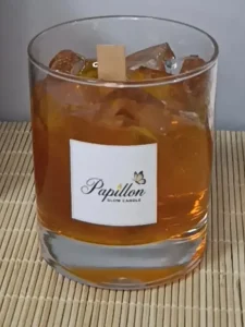 Viski bardağı mum: rocks bardağında amber renkli jel gövde, içinde şeffaf buz küpü efektli parçalar ve ortada ahşap fitil, ön yüzünde Papillon Glow etiketi