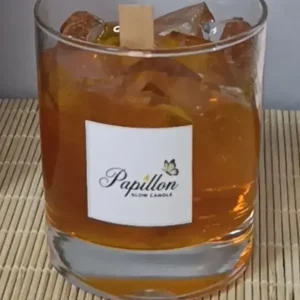 Viski bardağı mum: rocks bardağında amber renkli jel gövde, içinde şeffaf buz küpü efektli parçalar ve ortada ahşap fitil, ön yüzünde Papillon Glow etiketi