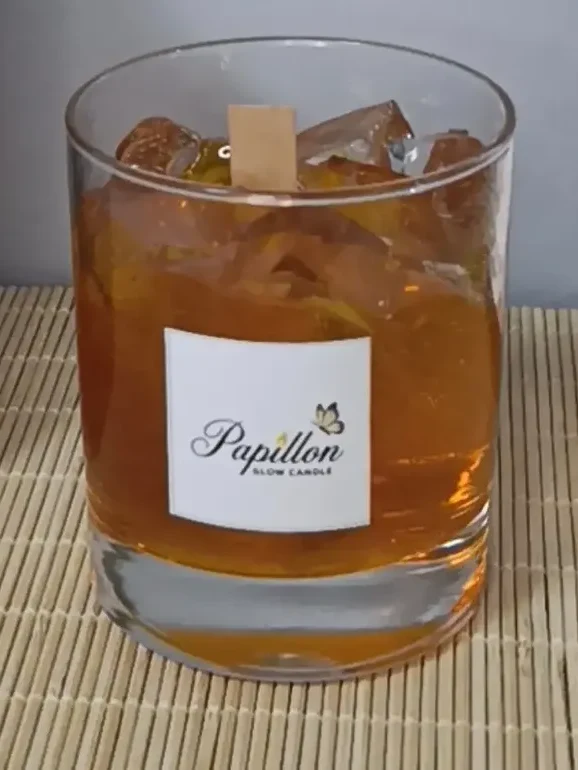 Viski bardağı mum: rocks bardağında amber renkli jel gövde, içinde şeffaf buz küpü efektli parçalar ve ortada ahşap fitil, ön yüzünde Papillon Glow etiketi